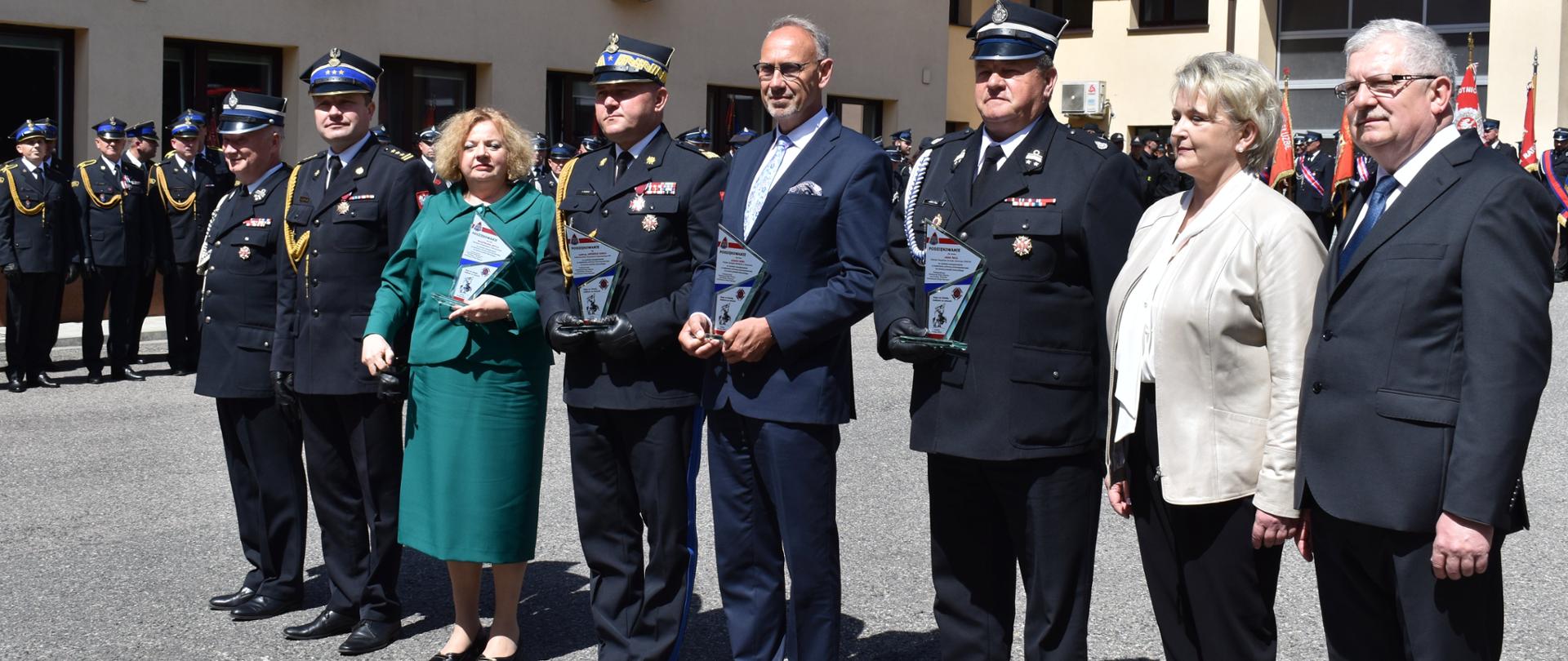 Zdjęcie zrobione na zewnątrz podczas uroczystości. Na zdjęciu, w szeregu stoją zaproszeni na uroczystość goście. Za nimi stoją ustawieni w szeregach funkcjonariusze Państwowej Straży Pożarnej. 