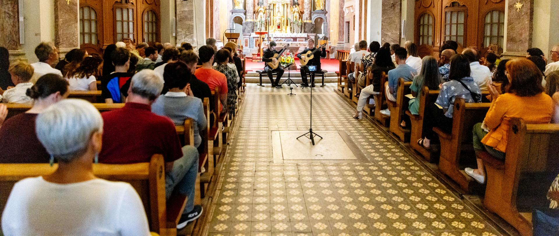 Wnętrze barokowego kościoła św. Szczepana w Postojnej wypełnione publicznością. Na tle ołtarza dwaj gitarzyści – Mak Grgić i Mateusz Kowalski – wykonują koncert, siedząc naprzeciw słuchaczy. Widok z końca nawy podkreśla liczną frekwencję i skupioną atmosferę wydarzenia.