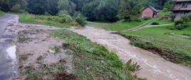Na zdjęciu widoczny rwący wezbrany potok, w tle dom mieszkalny.