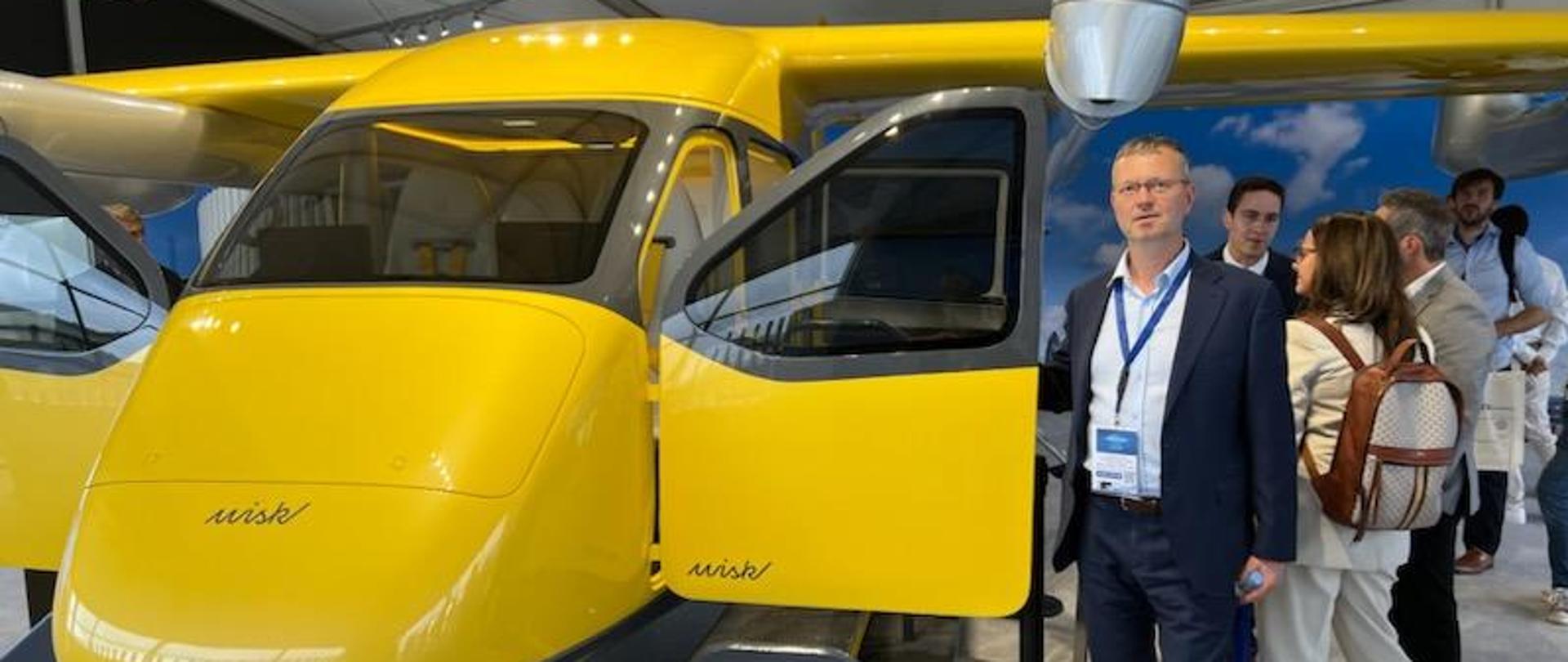 Wiceminister Jacek Bartmiński stoi przed żółtym samolotem. W tle inni zwiedzający targi. 