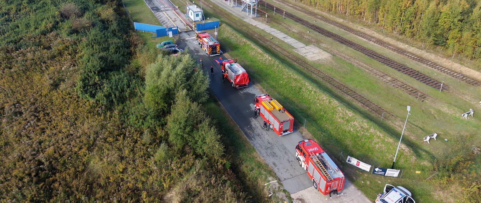 Na zdjęciu lotniczym widoczny jest teren zakładu przemysłowego z infrastrukturą kolejową oraz drogą dojazdową, na której ustawione są samochody pożarnicze i pojazd służb ratowniczych. W pobliżu znajdują się strażacy uczestniczący w ćwiczeniach. Teren zakładu otaczają rozległe obszary leśne i zielone.