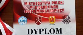Zdjęcie przedstawia pamiątkowy dyplom oraz brązowy medal za zajęcie III miejsca w mistrzostwach polski w kickboxingu służb mundurowych. Medal w kształcie trapezu w kolorze brązowym. U góry do medalu przypięta jest biało – czerwona wstążka służąca do zakładania medalu na szyję zawodnikowi podczas ceremonii dekoracji. Dyplom w kolorze czerwono – białym patrząc od góry. Na dyplomie widnieje napis (od góry): mistrzostwa polski w kickboxingu służb mundurowych, poniżej umieszczone są loga partnerów oraz znajduje się napis: dyplom dla szymona grabowskiego za zajęcie trzeciego miejsca na mistrzostwach polski służb mundurowych. Poniżej określona jest kategoria wagowa do 89 kg. Na samym dole: Żyrardów, 23 - 25 września 2022 r. 