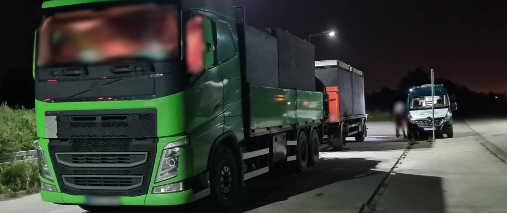 Na pierwszym planie (od lewej strony): przód kabiny i lewa strona samochodu ciężarowego z szambami betonowymi. W tle (od prawej strony): załoga kierowców przeładowanej ciężarówki stoi obok oznakowanego furgonu mazowieckiej ITD. 