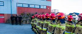 Zdjęcie przedstawia strażaków JRG Sanok podczas oficjalnej zbiórki z okazji NŚN. Strażacy stoją w dwuszeregu. W tle poczet flagowy i budynek KP PSP Sanok