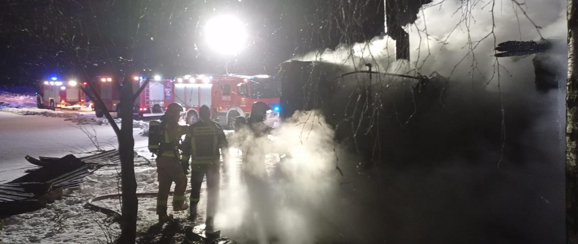 Na zdjęciu widzimy palący się budynek dużo dymu 3 strażaków rosnące dwa drzewa na ziemi zalegający śnieg a w tle trzy samochody strażackie