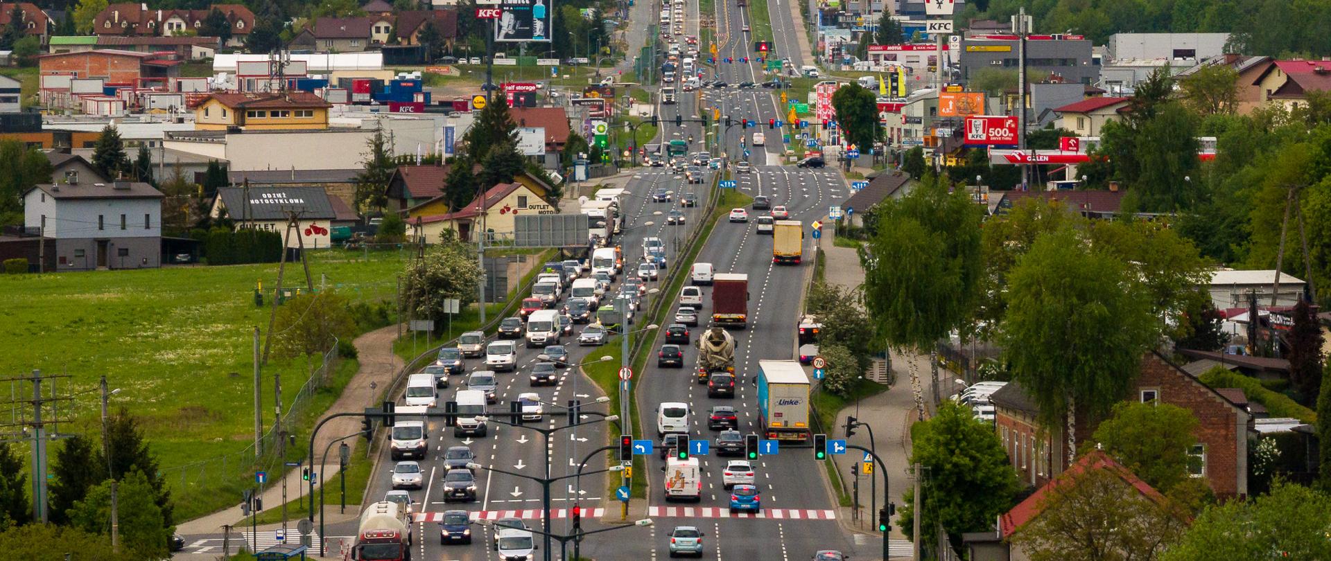 Widok na szeroką, wielopasmową drogę miejską z dużym natężeniem ruchu, otoczoną zabudową usługową i mieszkaniową, z zielenią w tle oraz pagórkowatym krajobrazem.
