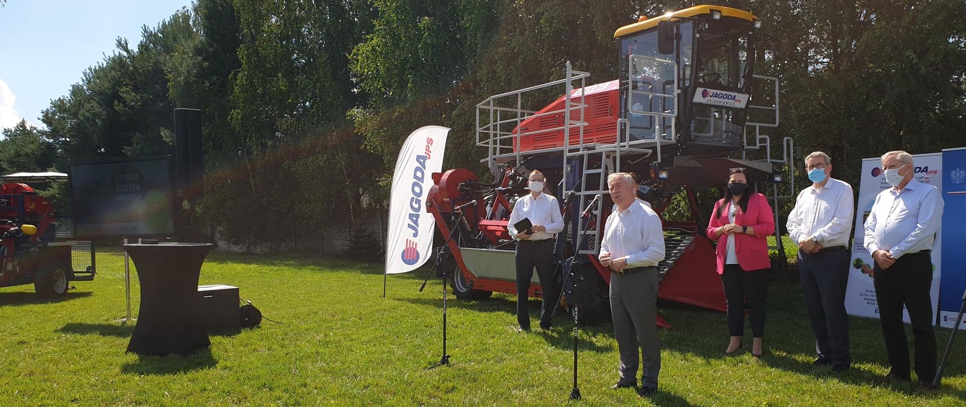w plenerze na trawie widoczne sprzęty rolnicze. w centralnej części na tle linii drzew widoczny kombajn do zbioru owoców jagodowych. obok stoją :