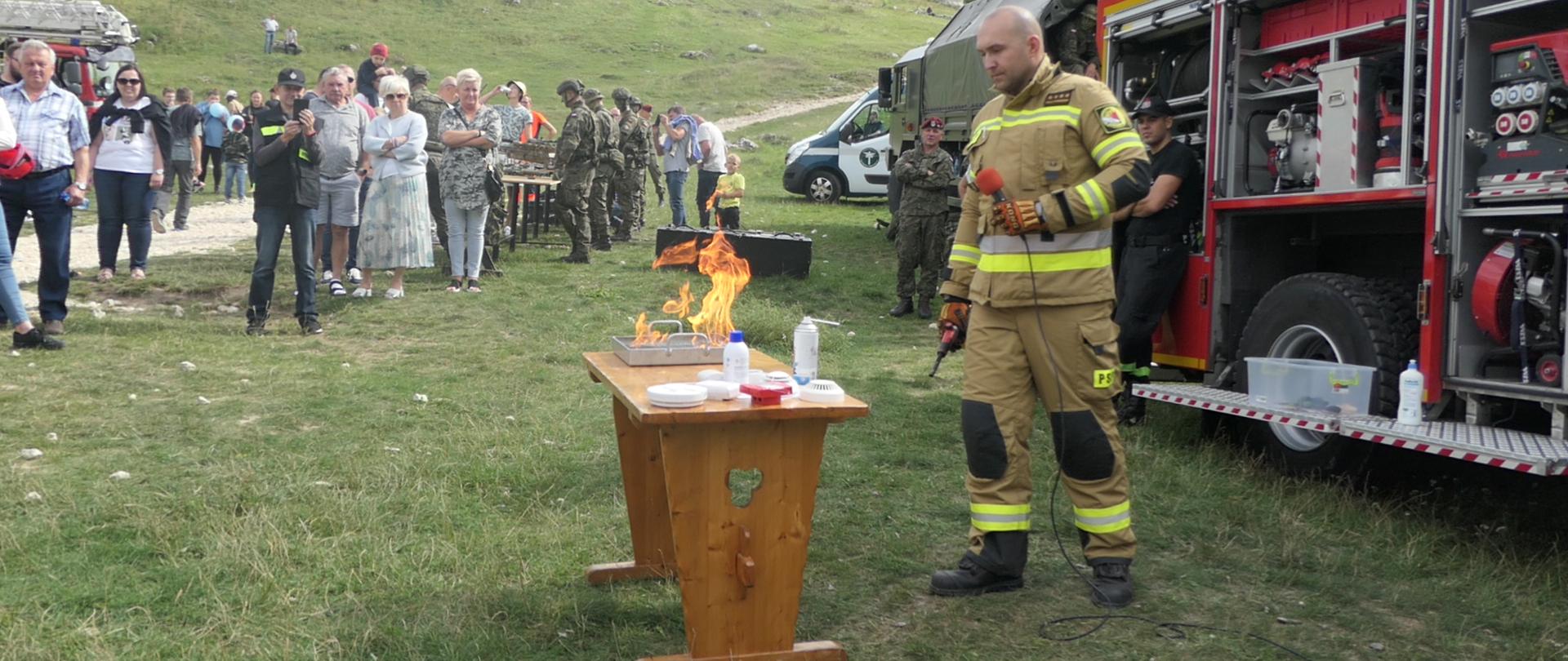 Na pierwszym planie biurko, na którym leżą czujki dymu oraz metalowy pojemnik z substancją która płonie. Po prawej stronie strażak w umundurowaniu bojowym, trzymający w ręku mikrofon, objaśniający przeprowadzane doświadczenie. W głębi osoby obserwujące pokaz
