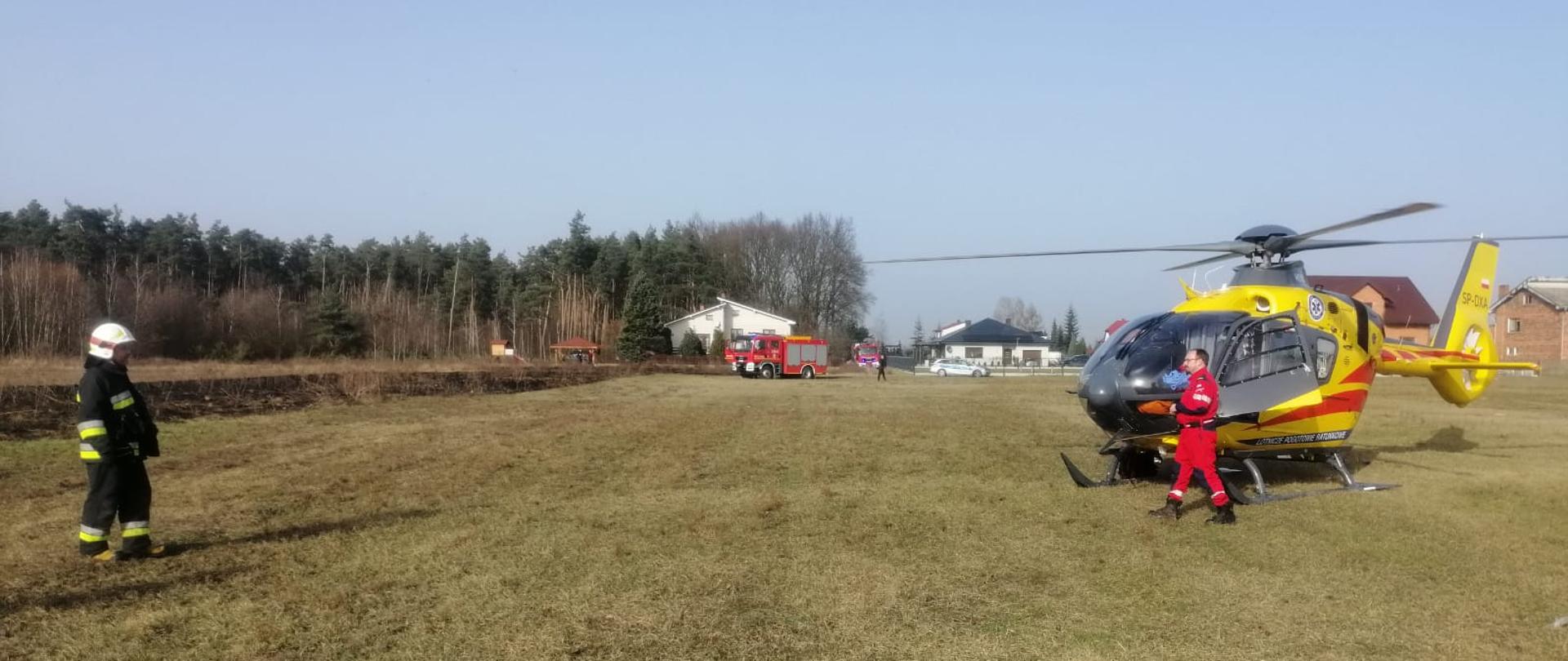 Na zdjęciu widać helikopter Lotniczego Pogotowia Ratunkowego w kolorze żółtym, Obok można zauważyć ratownika medycznego ubranego w pomarańczowy kombinezon oraz strażaka w mundurze koloru czarnego z widocznymi elementami odblaskowymi. W tle widać samochody strażackie w kolorze czerwonym oraz spaloną powierzchnię trawy. Z prawej strony z tyłu znajdują się zabudowania oraz jest widoczny radiowóz Policji.
