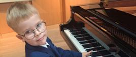 Na zdjęciu chłopiec, blondyn w okularach i granatowym garniturze. Chłopiec gra na czarnym fortepianie.Tło obrazka w kolorze jasno brązowym.