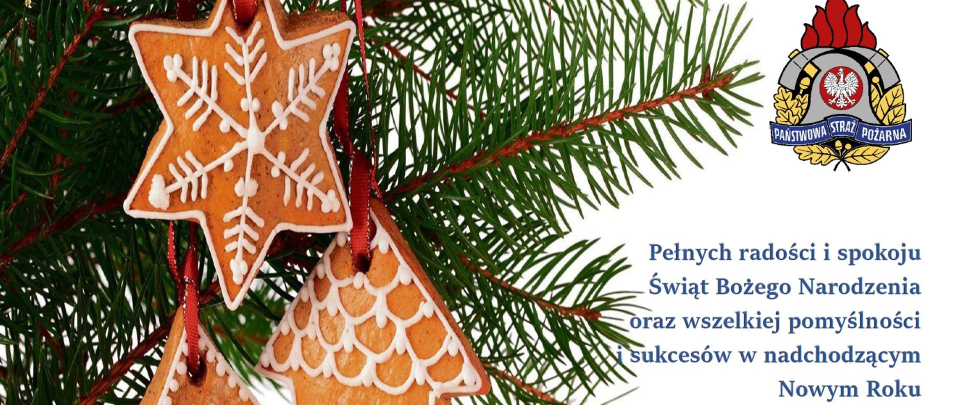 Zielone gałązki a na nich zawieszone pierniki w kształcie gwiazdki, choinki i domku. Po prawej stronie kolorowy znak PSP i życzenia Komendanta Powiatowego.