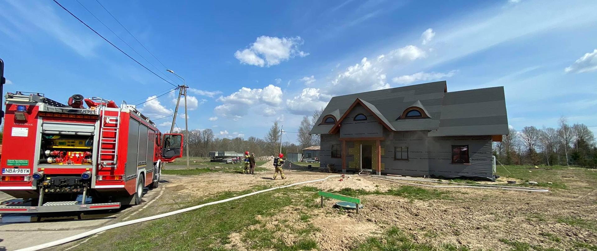 Po lewej stronie widać czerwony wóź bojowy. Obok widać strażaków i osobę postronną oraz dom, w którym doszło do pożaru