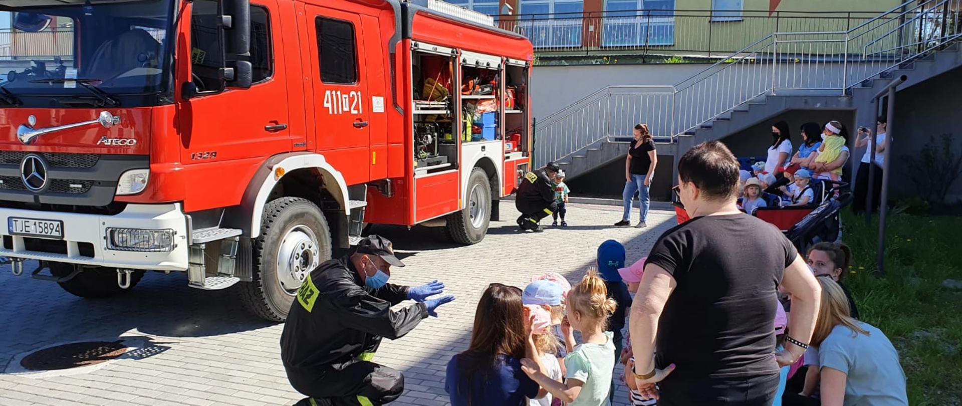 Zdjęcie przedstawia kilkanaście małych dzieci pod opieką nauczycieli. Jeden ze strażaków w przykucniętej pozycji przemawia do dzieci oraz gestykuluje. Ma założoną maseczkę zakrywającą usta i nos oraz rękawiczki lateksowe. Po lewej stronie znajduje się samochód ratowniczo- gaśniczy z widocznym sprzętem pożarniczym.
