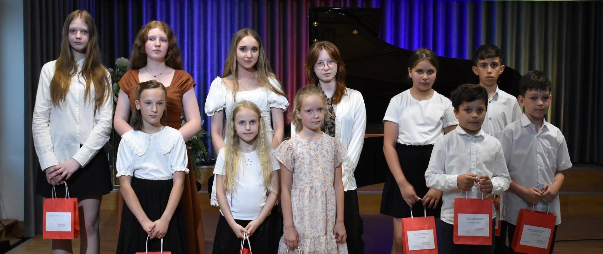 Grupa dzieci i młodzieży pozuje na scenie przed fortepianem w Państwowej Szkole Muzycznej I stopnia im. Witolda Lutosławskiego w Jaśle; część osób trzyma czerwone torby z upominkami.