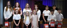 Grupa dzieci i młodzieży pozuje na scenie przed fortepianem w Państwowej Szkole Muzycznej I stopnia im. Witolda Lutosławskiego w Jaśle; część osób trzyma czerwone torby z upominkami.