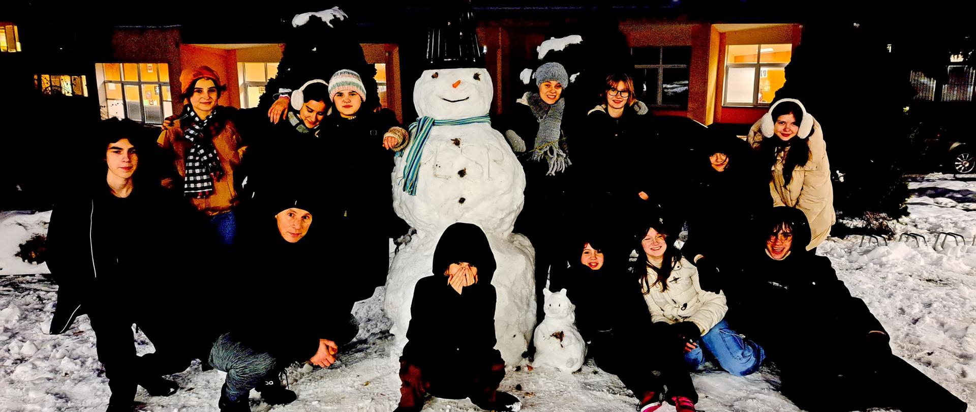 Zdjęcie grupowe przedstawia uczniów i nauczyciela Tomasza Krasowskiego. Pozują oni do zdjęcia. Stoją wokół śnieżnego bałwana na tle budynków Ogólnokształcącej Szkoły Muzycznej I i II st. im. Karola Lipińskiego w Lublinie.