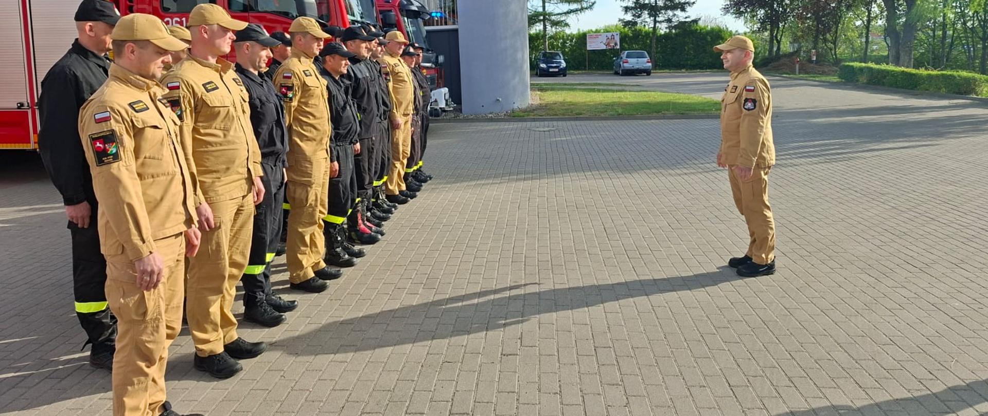 Wielkopolski Komendant Wojewódzki wizytował konińskich strażaków. Komendant składa życzenia na placu przed budynkiem. Strażacy stoją w szyku za nimi samochody strażackie