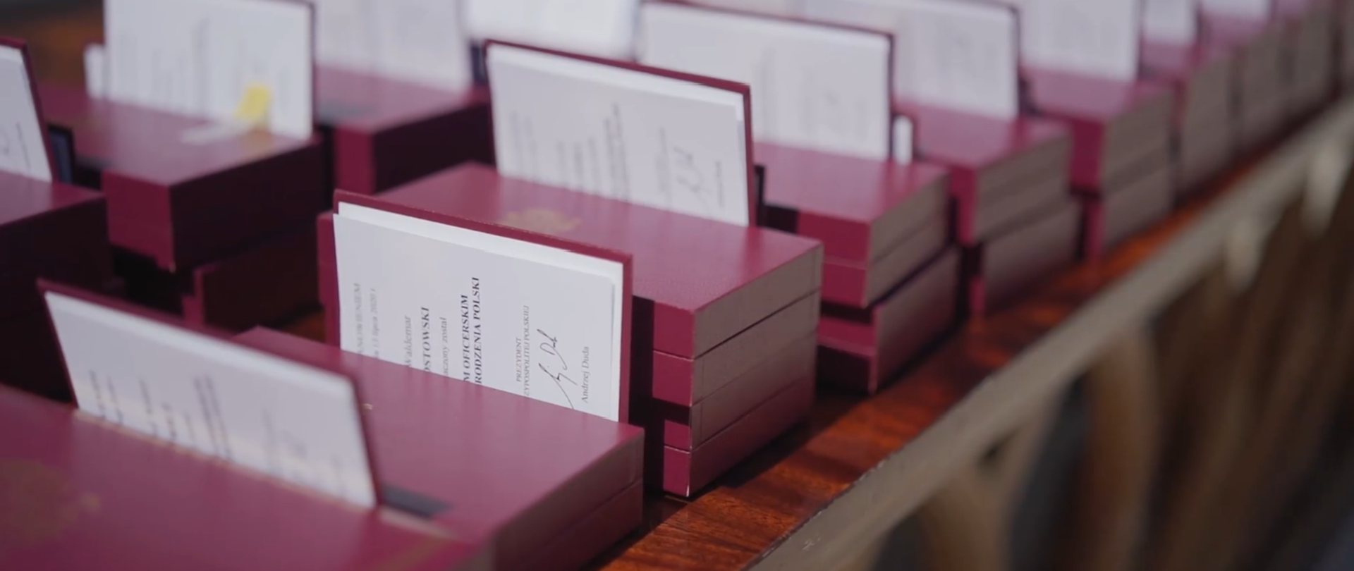 Zdjęcie przedstawia ułożone na stole bordowe pudełeczka, w których znajdują się odznaczenia państwowe oraz listy gratulacyjne w formie książeczek.