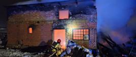 Murowany z czerwonej cegły budynek objęty pożarem. Strażak podaje prąd wody do wnętrza budynku. Zadymienie wydobywa się z dachu.