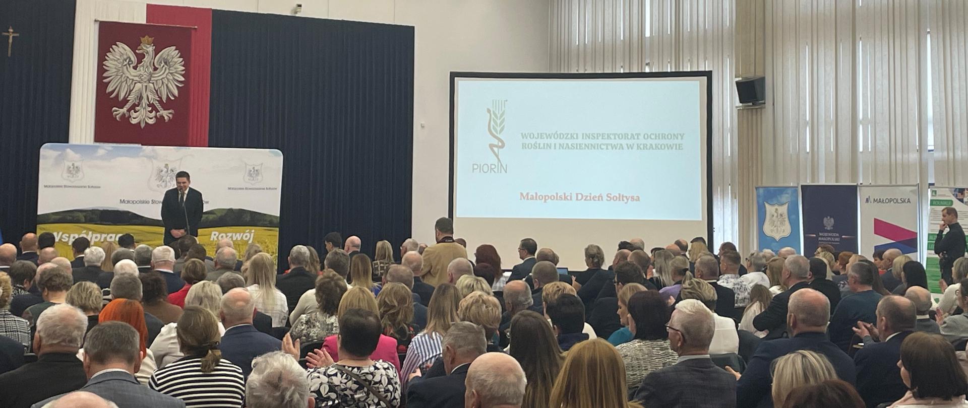 Widok na salę konferencyjną i uczestników spotkania, na drugim planie widoczna scena, a na niej osoba przemawiająca (mężczyzna), z prawej strony widok prezentacji multimedialnej