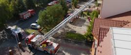 Widok z dronu na teren działań przed szkoła, na pierwszym planie drabina mechaniczna i strażak w koszu przygotowujący się do wejścia do budynku