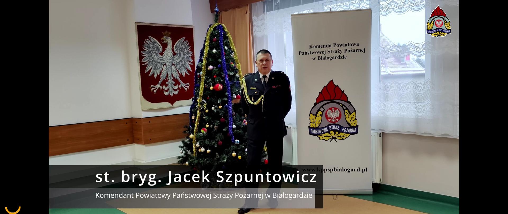Zdjęcie przedstawia stojącego Komendanta PSP. Po jego lewej stronie na ścianie wisi godło Polski a po prawej baner KP PSP Białogard. Tło stanowi duża, zielona choinka. Komendant jest ubrany w mundur wyjściowy