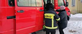 Strażak pomaga osobie starszej wsiąść do samochodu strażackiego podtrzymując ją za rękę. Kobieta drugą ręką pomaga sobie chwytając pojazdu.
