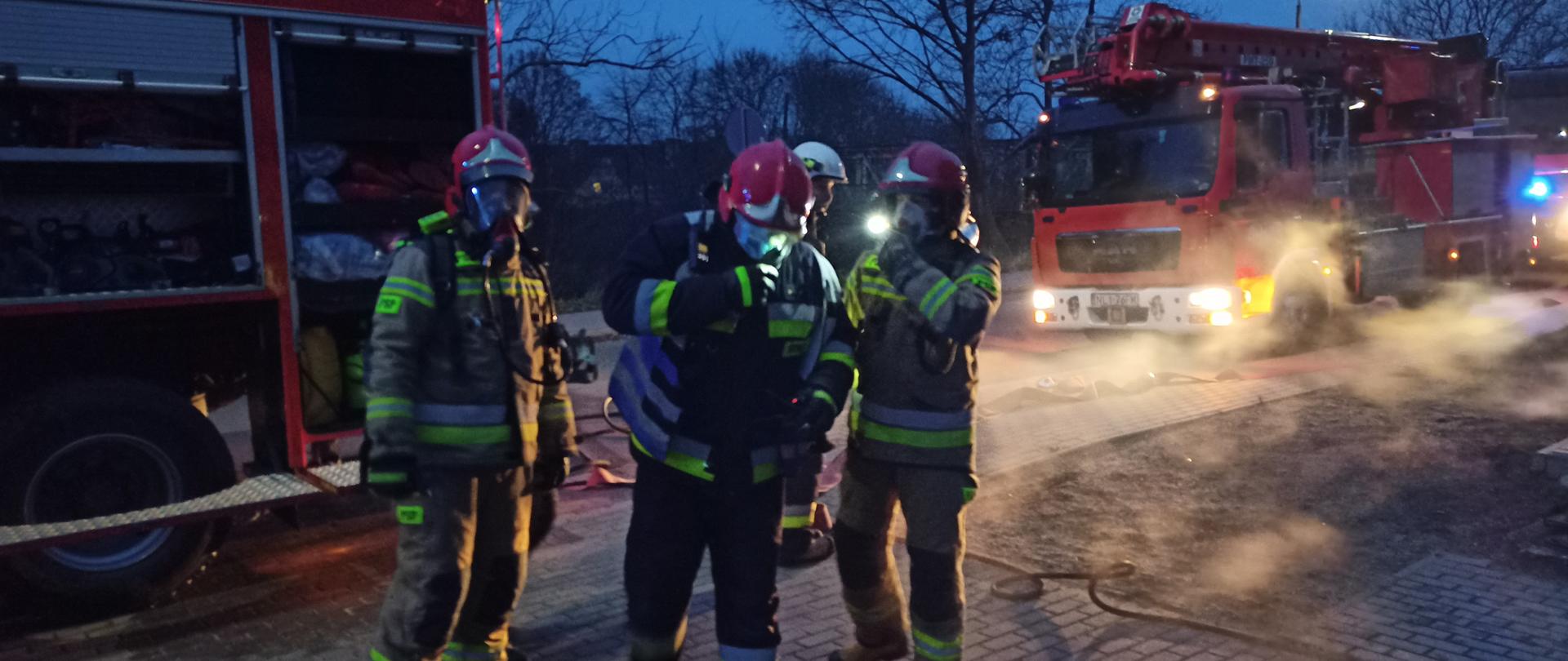 Na pierwszym planie widoczny jest Kierujący Działaniem Ratowniczym, który prowadzi korespondencje radiową z stanowiskiem kierowania Komendanta Powiatowego w Lidzbarku Warmińskim. Obok niego stoi dwóch ratowników PSP, jeden z nich ubrany sprzęt ochrony dróg oddechowych a za nimi idzie strażak OSP. W drugim planie znajdują się trzy samochody pożarnicze.