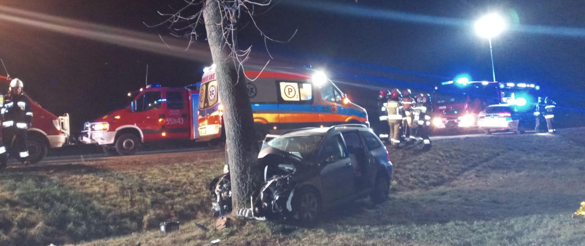 Zdjęcie przedstawia samochód osobowy po uderzeniu przodem w drzewo, w tle widać trzy wozy bojowe straży pożarnej, karetkę pogotowia, radiowóz policyjny oraz strażaków w ubraniach bojowych