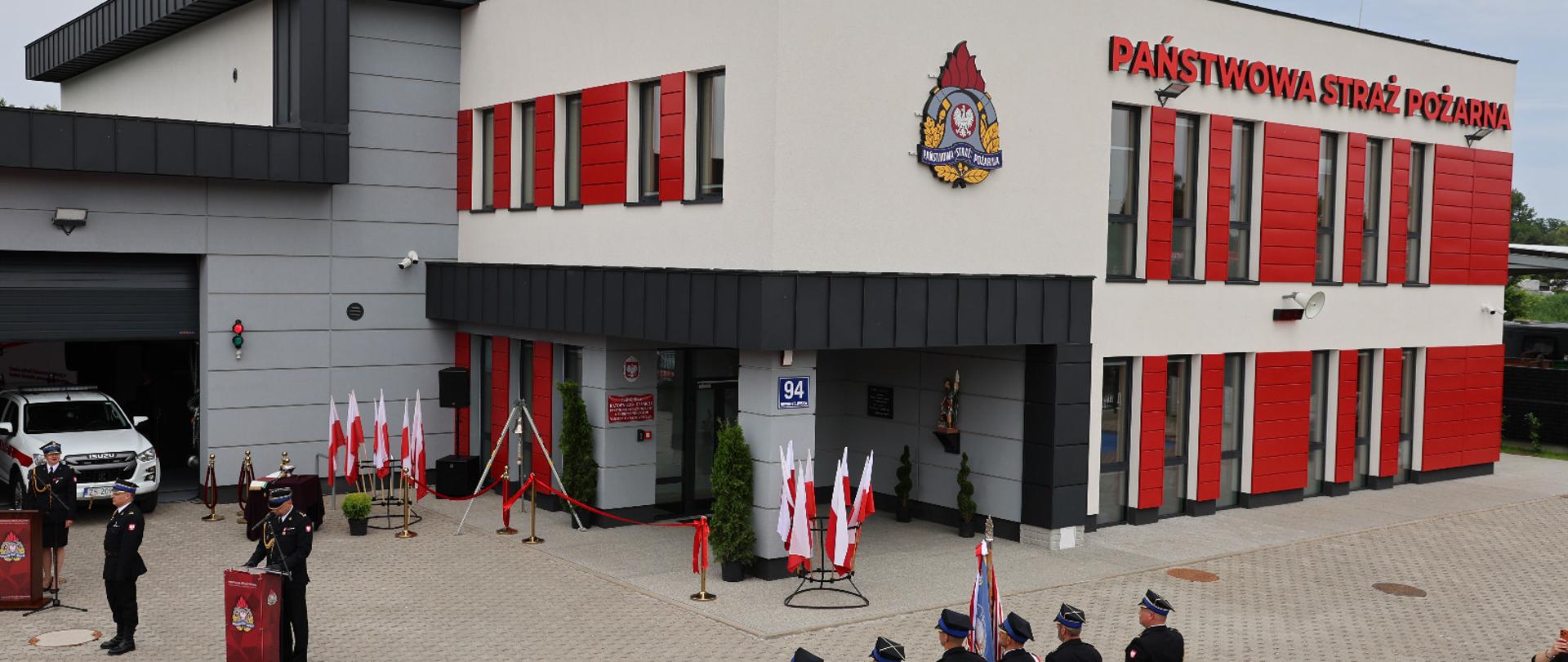 Uroczystość przed budynkiem Państwowej Straży Pożarnej. Przed wejściem ustawieni są funkcjonariusze PSP i zaproszeni goście, a przy mównicy przemawia strażak w mundurze wyjściowym. Na placu widoczne są poczty sztandarowe, biało-czerwone flagi i oznaczenia PSP na elewacji budynku.