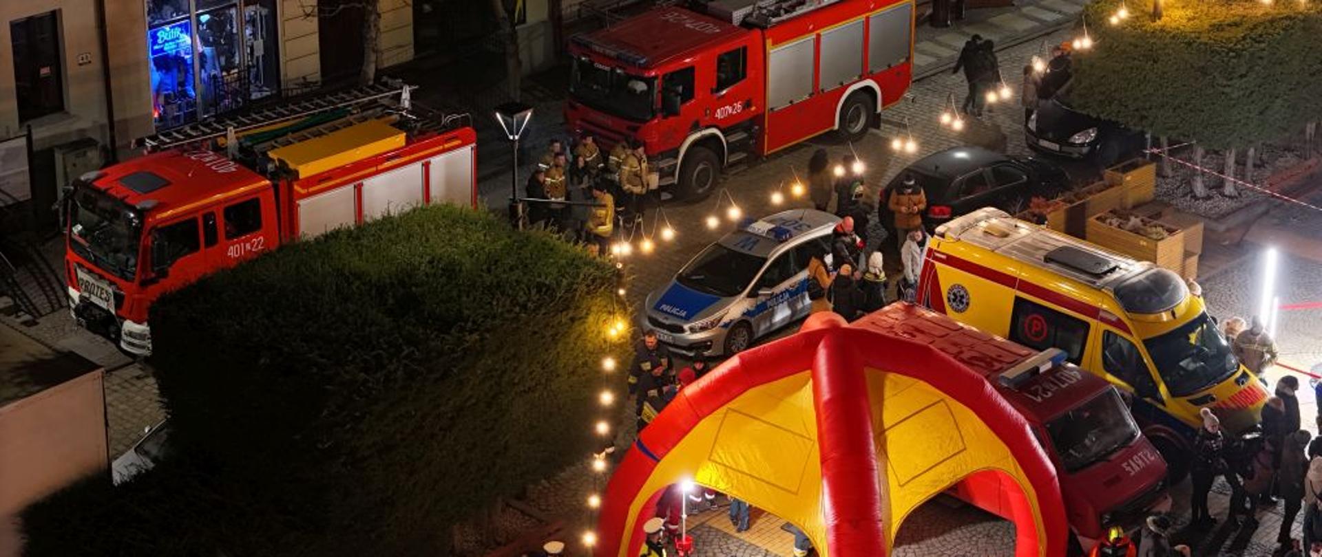 Widok z drona na strażaków i wozy gaśnicze. Strażacy stoją przy pojeździe w piaskowych mundurach. Zdjęcie wykonane w nocy po zmierzchu.