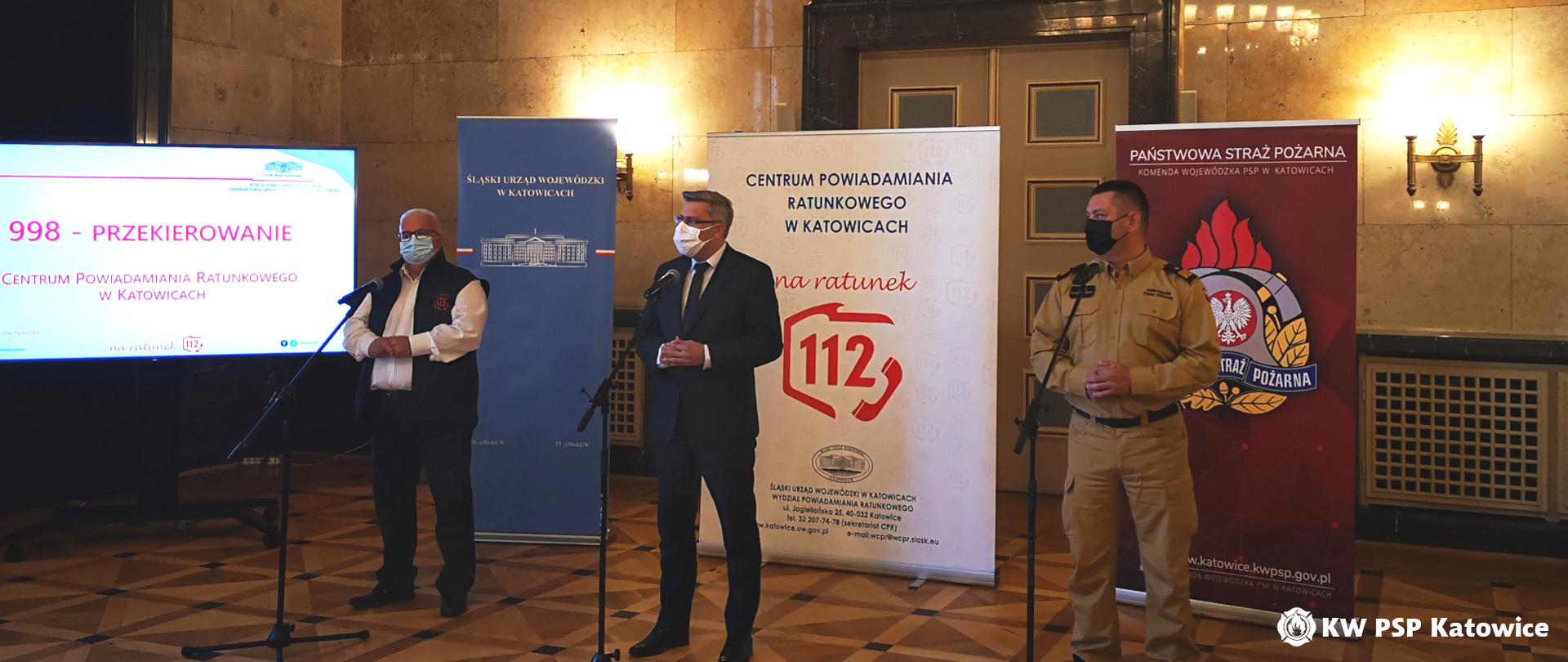 Na zdjęciu widoczna jest sala marmurowa w Śląskim Urzędzie Wojewódzkim. Na tle ściany ustawione są od prawej banery Komendy Wojewódzkiej PSP w Katowicach, Śląskiego Urzędu Wojewódzkiego oraz Centrum Powiadamiania Ratunkowego. Na ich tle, przed mikrofonami stoją od strony prawej: śląski komendant wojewódzki PSP ,wojewoda śląski, dyrektor CPR w Katowicach. Po lewej stronie na ekranie telewizora wyświetla się prezentacja. 