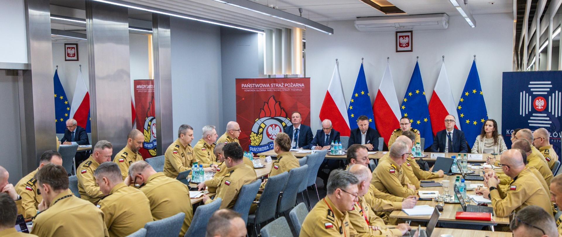 Szerokie ujęcie sali konferencyjnej w Komendzie Głównej PSP; liczni funkcjonariusze siedzą przy stołach i uczestniczą w naradzie.