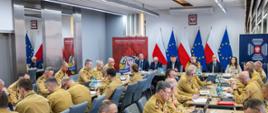 Szerokie ujęcie sali konferencyjnej w Komendzie Głównej PSP; liczni funkcjonariusze siedzą przy stołach i uczestniczą w naradzie.