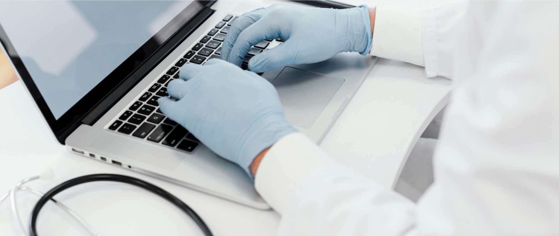 Osoba pracująca na laptopie (widoczne tylko ręce) ma na dłoniach rękawiczki oraz ubrana jest w fartuch laboratoryjny co może stwierdzać że jest to pracownik labaratorium.