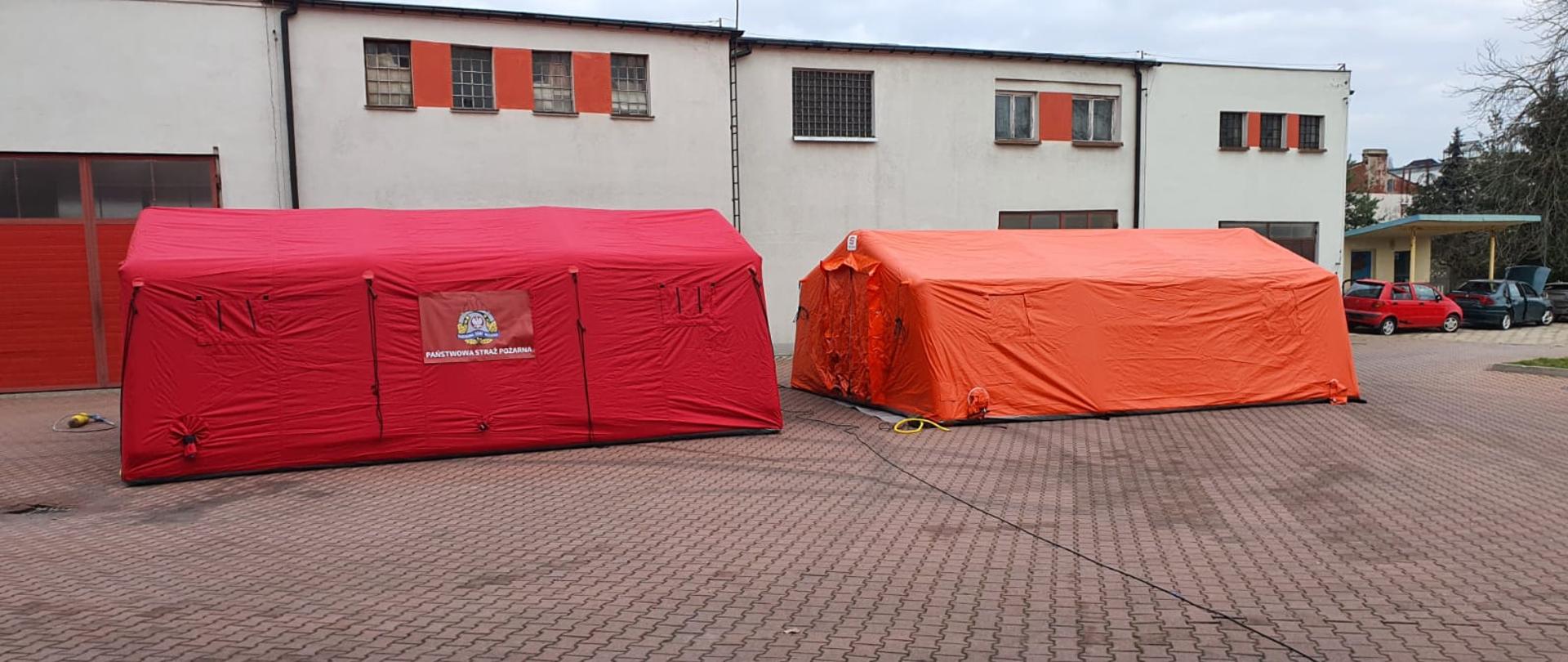 Na zdjęciu widzimy dwa rozłożone namioty ratownicze. Z lewej strony kadru widzimy namiot koloru czerwonego z naniesionym logo Państwowej Straży Pożarnej.
Z prawej strony kadru widzimy namiot koloru pomarańczowego. Namioty rozłożone są na placu manewrowym który jest utwardzony kostką brukową koloru czerwonego. W tle widzimy budynki gospodarcze koloru kremowego. Z prawej strony kadru w tle widzimy dwa samochody koloru czerwonego i ciemno zielonego stojących pod wiatą. 
