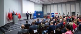 
Grupa ludzi w sali konferencyjnej, 3 osoby stojące na scenie
