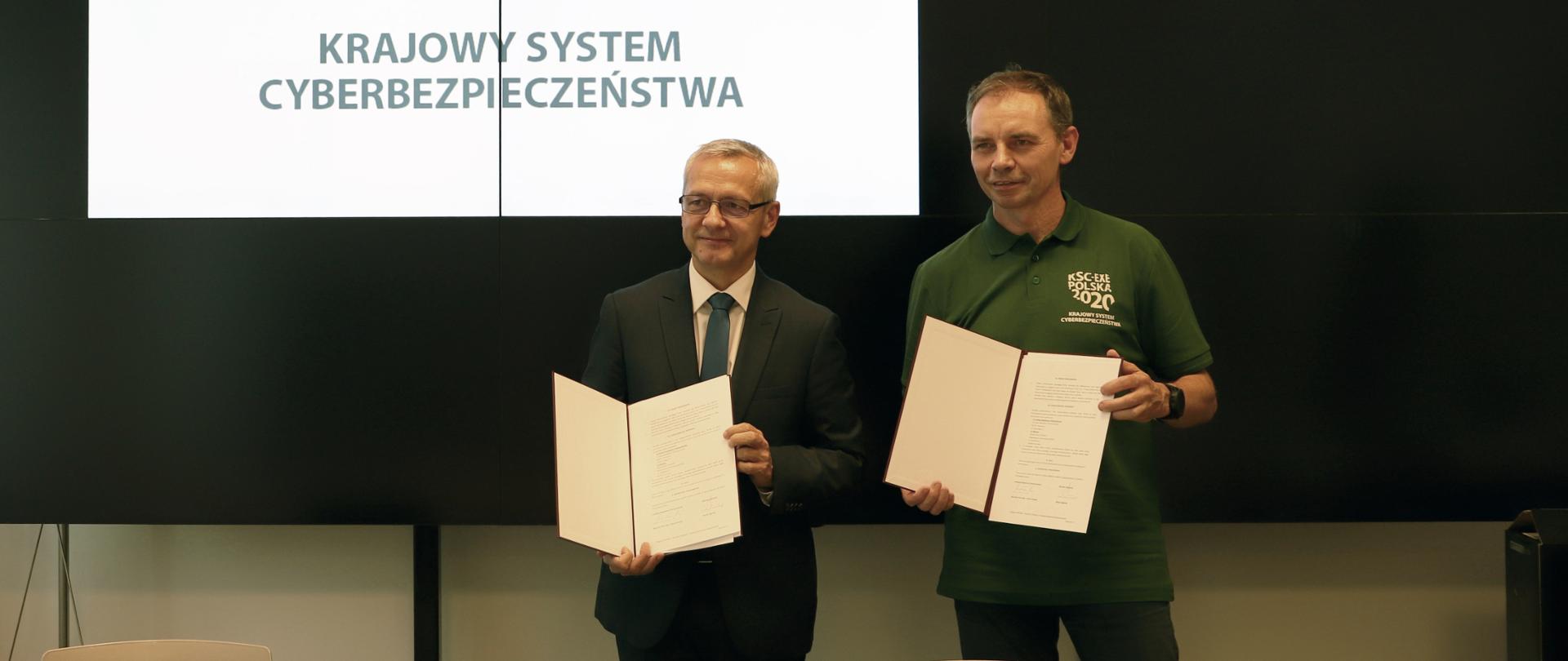 Minister cyfryzacji Marek Zagórski i Mirosław Maj, prezes Fundacji Bezpieczna Cyberprzestrzeń pozują do zdjęcia z teczkami, w których jest podpisane przez obie strony porozumienie.