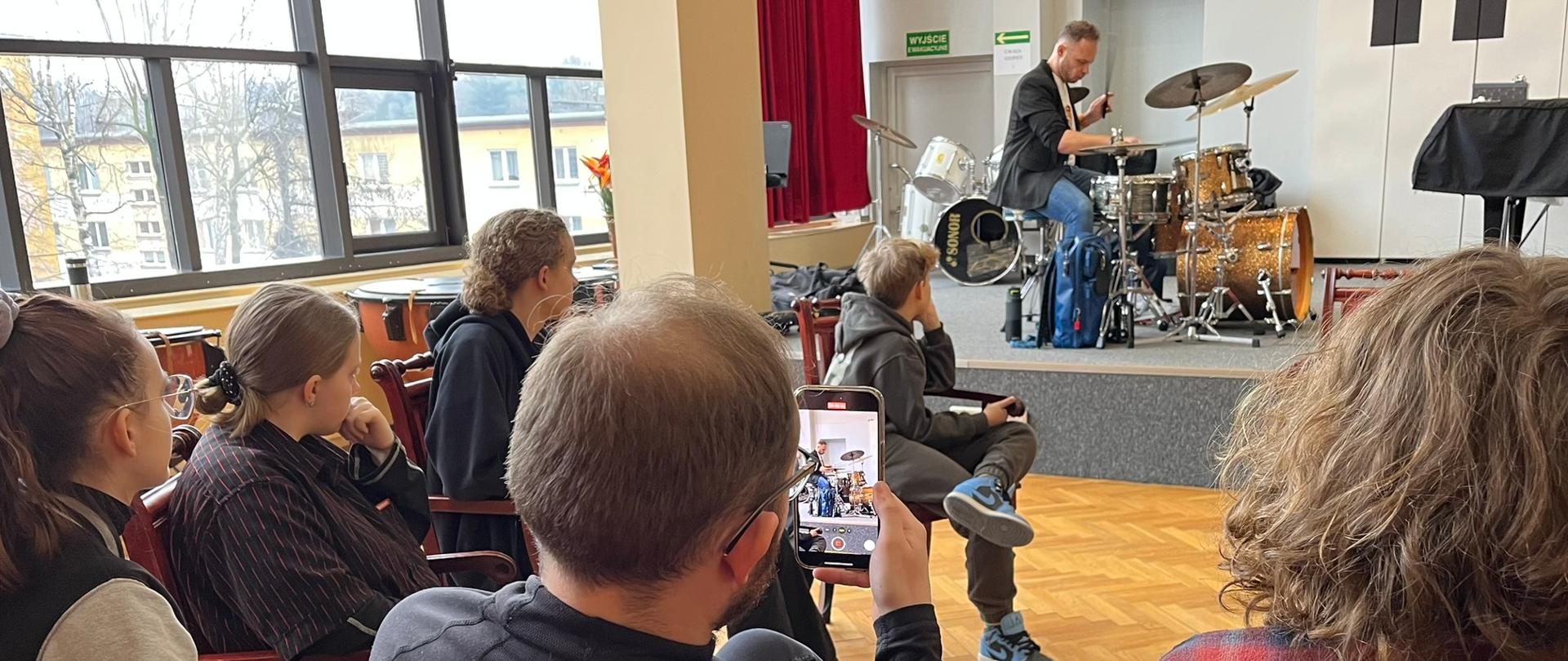 Mężczyzna gra na zestawie perkusyjnym, z przodu widoczna od tyłu publiczność.