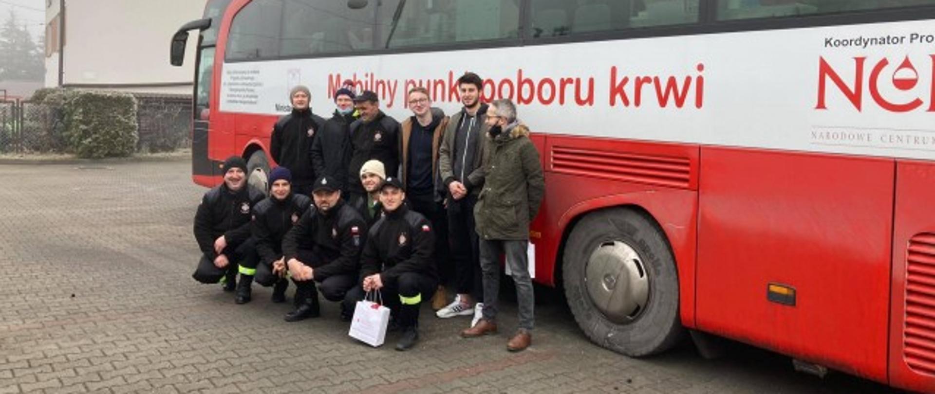 11 osób w dwóch rzędach pozuje do pamiątkowego zdjęcia z akcji Krwiodawstwa na tle autobusu pełniącego funkcję mobilnego punktu poboru krwi