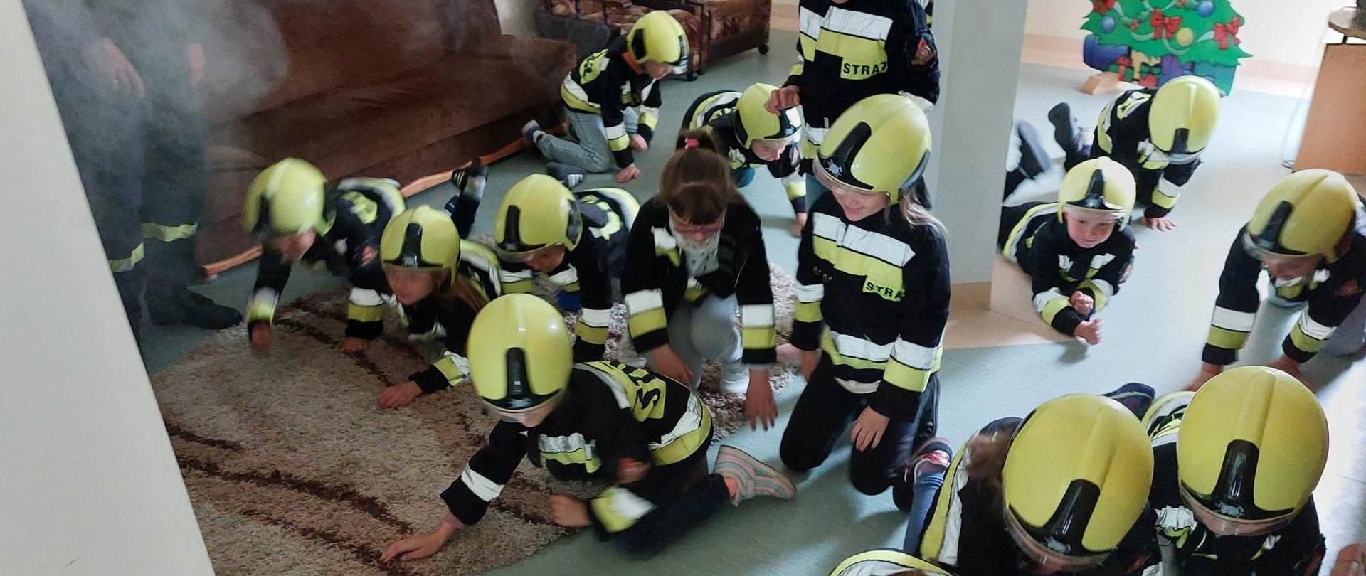 Dzieci wychodzące na czworaka z zadymionego pomieszczenia, ubrane w żółte hełmy oraz kurtki ubrania specjalnego,. 