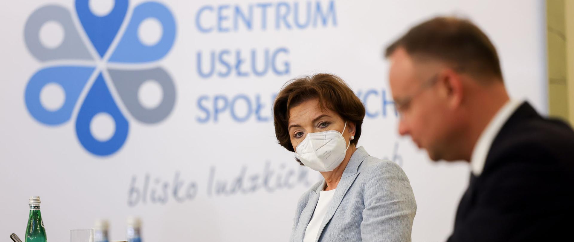 Zbliżenie na postać minister Maląg. Na pierwszym planie dość niewyraźna postać prezydenta Dudy. Oboje siedzą przy stole. Na którym widać butelki z wodą, szklankę, złożone okulary.