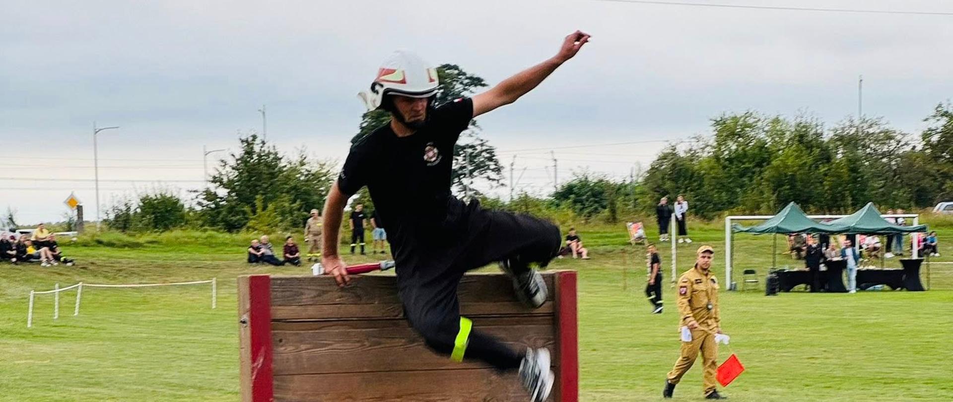 Strażak ochotnik w czarnym stroju i białym hełmie przeskakuje przez drewnianą przeszkodę podczas zawodów sportowo-pożarniczych. W tle widoczni są sędzia z czerwoną chorągiewką oraz kibice i namioty organizatorów na zielonym boisku. Pogoda jest pochmurna, a atmosfera sportowa i zaangażowana.