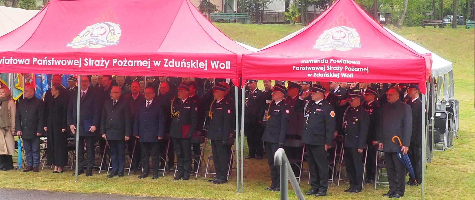 strażacy w mundurach wyjściowych i inni uczestnicy uroczystości stoją pod namiotem bankietowym, namioty z logo PSP i napisami, strażacy salutują