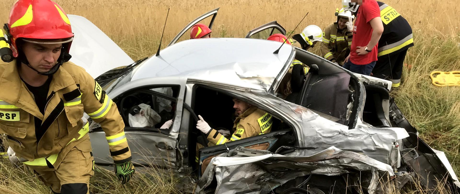 Srebrny samochód znajduje się w rowie na pierwszym planie strażak wychodzi z rowu i idzie natomiast przy samochodzie widać innych strażaków którzy udzielają pierwszej pomocy poszkodowanej jeden strażak znajduje się w samochodzie w pobliżu stoi mężczyzna w czerwonej koszulce. W oddali pole na którym rośnie zborze