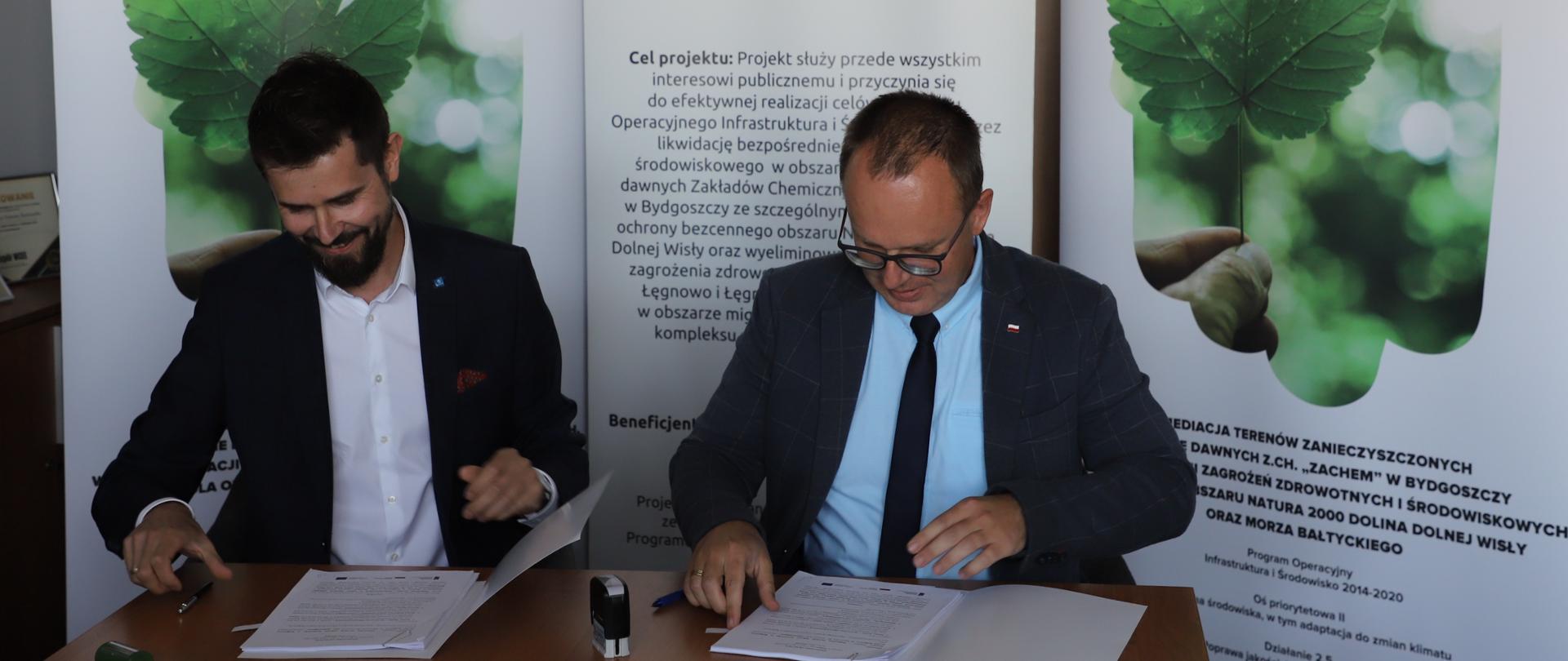 Dwóch mężczyzn siedzi przy stole, przed nimi dwa egzemplarze umowy, długopisy i pieczątki