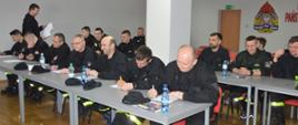 Egzamin końcowy. Świetlica Komendy Powiatowej PSP w Rawiczu. Przy stołach siedzą egzaminowani strażacy. Przewodniczący komisji egzaminacyjnej rozdaje im testy wraz z kartami odpowiedzi. 