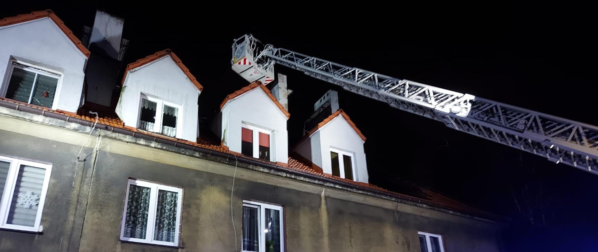 Samochód strażacki z wysuniętą drabiną mechaniczną oświetla z kosza poddasze wewnątrz, którego strażacy prowadzą prace rozbiórkowe.
