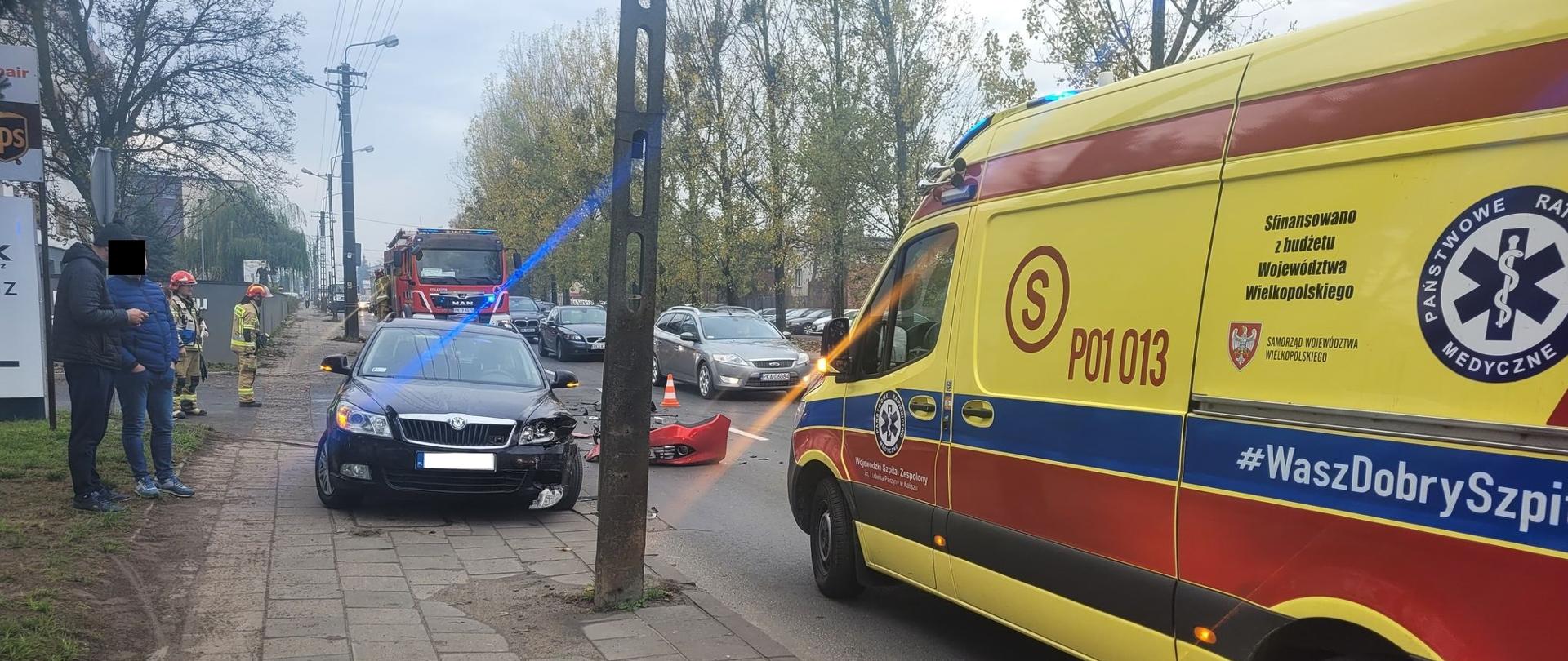 Zdjęcie wykonane w ciągu dnia. Widać pojazdy służb, a obok osoby postronne oraz funkcjonariuszy. Zdarzenie ma miejsce na drodze publicznej. Inni uczestnicy ruchu. Zachmurzenie duże.