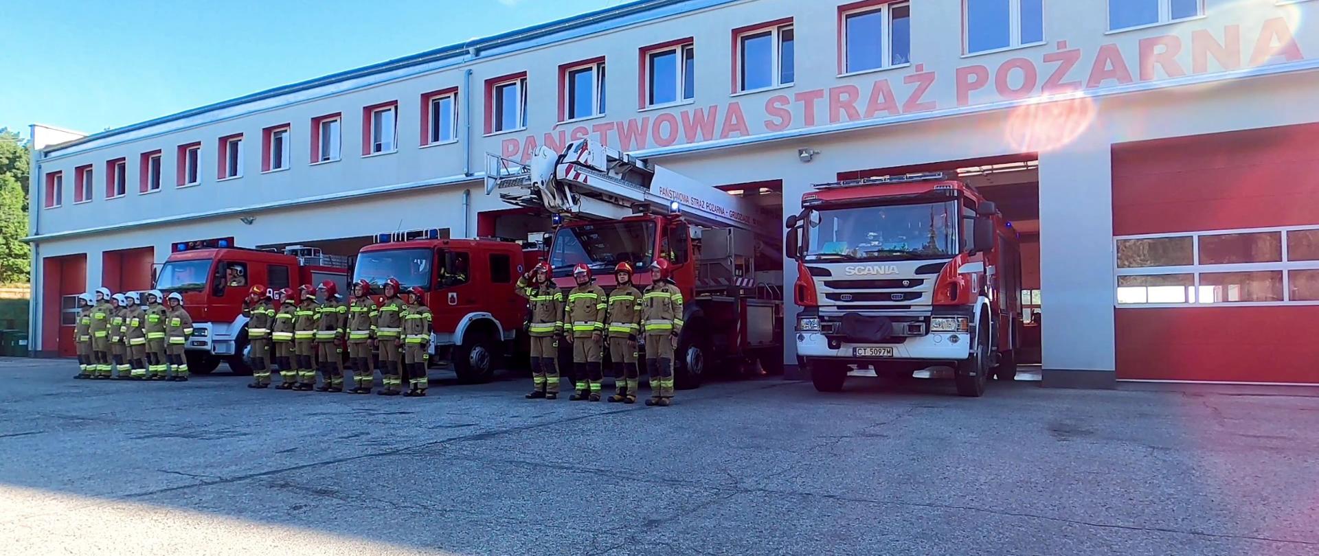 Zdjęcie przedstawia 4 czerwone, ciężarowe, strażackie samochody, przed którymi w trzech szeregach stoją strażacy ubrani w piaskowych mundurach i czerwonych oraz białych hełmach. Dowódcy oddają honor. Pojazdy w połowie stoją na utwardzonym placu, a w połowie w garażu budynku z napisem „Państwowa Straż Pożarna”